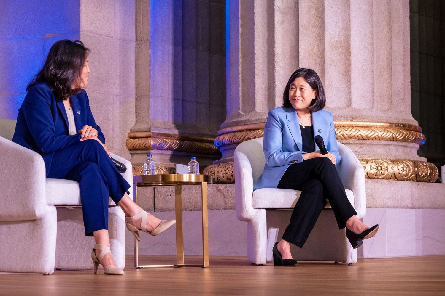Two women engage in a panel discussion seated in white chairs at a formal event venue.