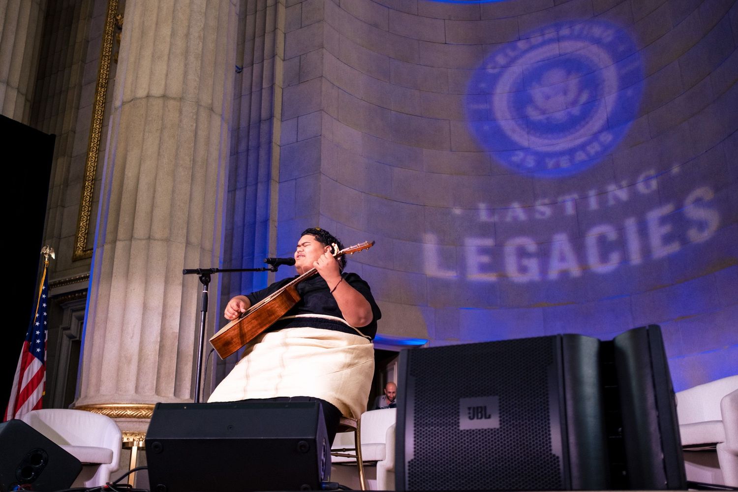 Musician performs on stage at Lasting Legacies celebration event beneath a glowing government seal projection.