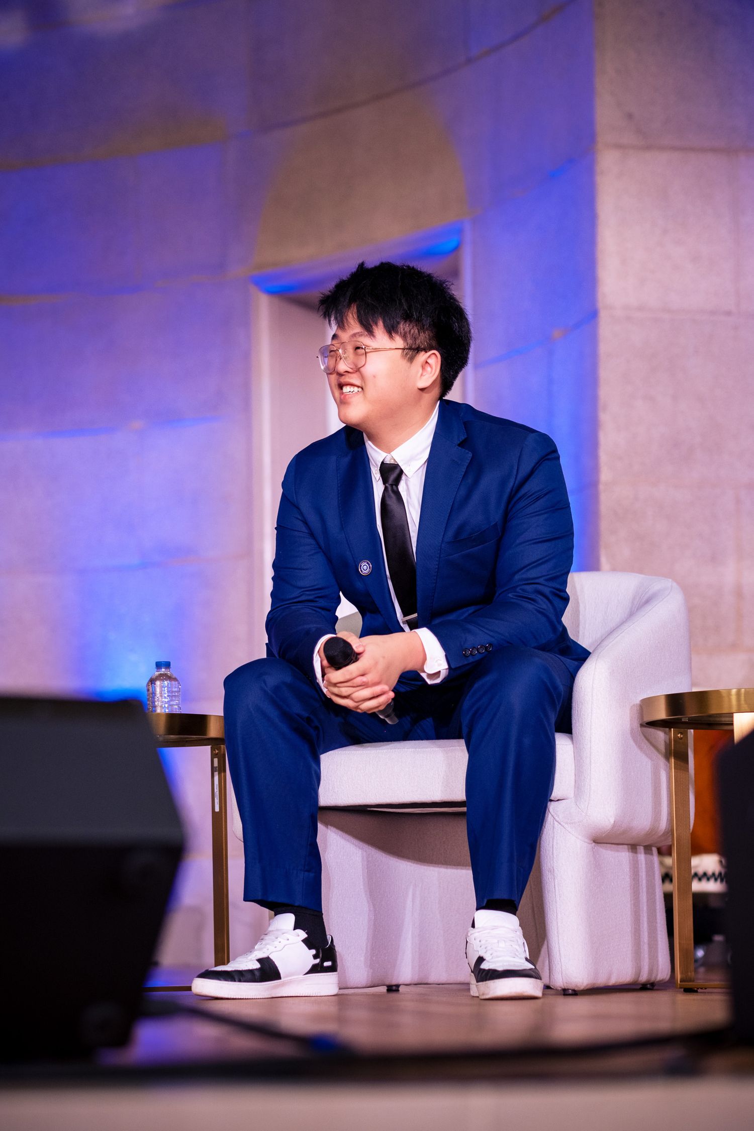 Young man in blue suit and glasses sits smiling on white chair at formal event panel discussion.