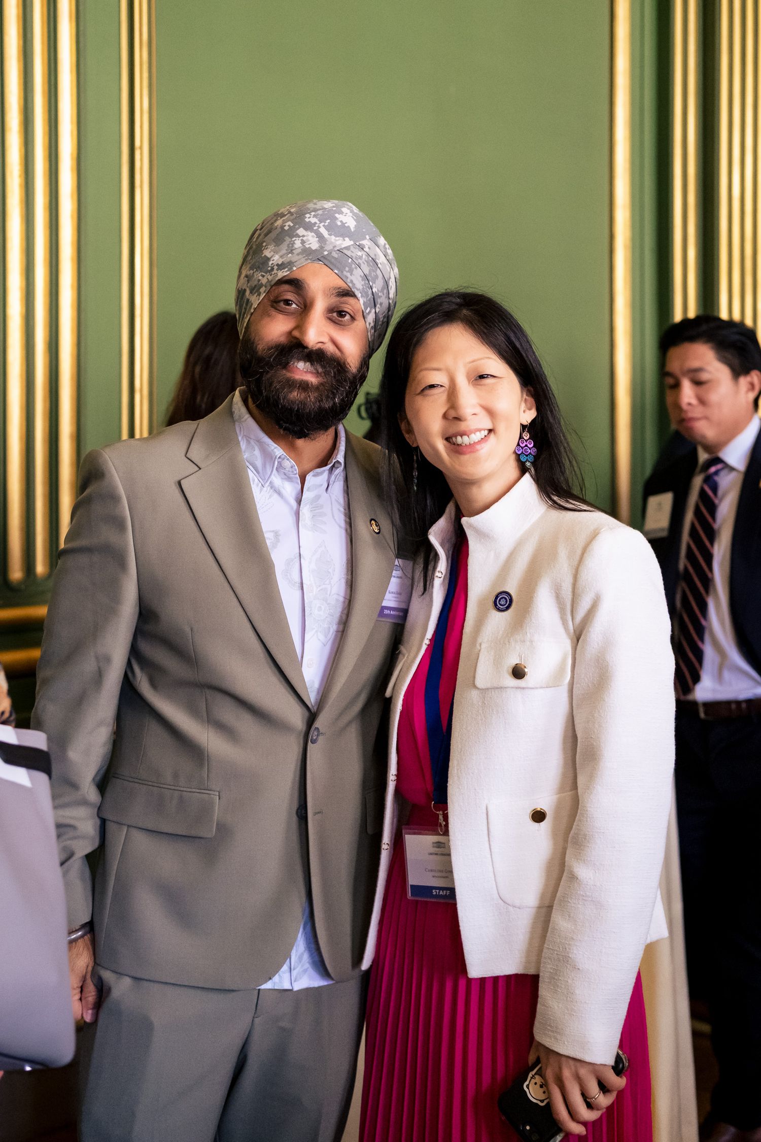 Two professionals smile together at a formal event, man in turban and suit, woman in white blazer and pink skirt.