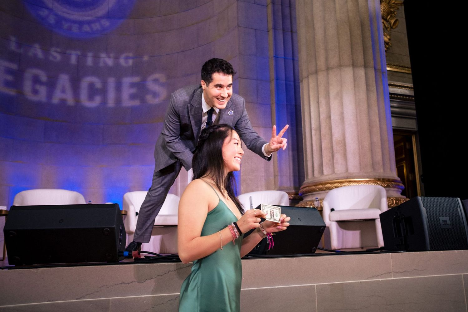 Man on stage gestures toward a woman in a green dress holding an award at a Lasting Legacies gala event.