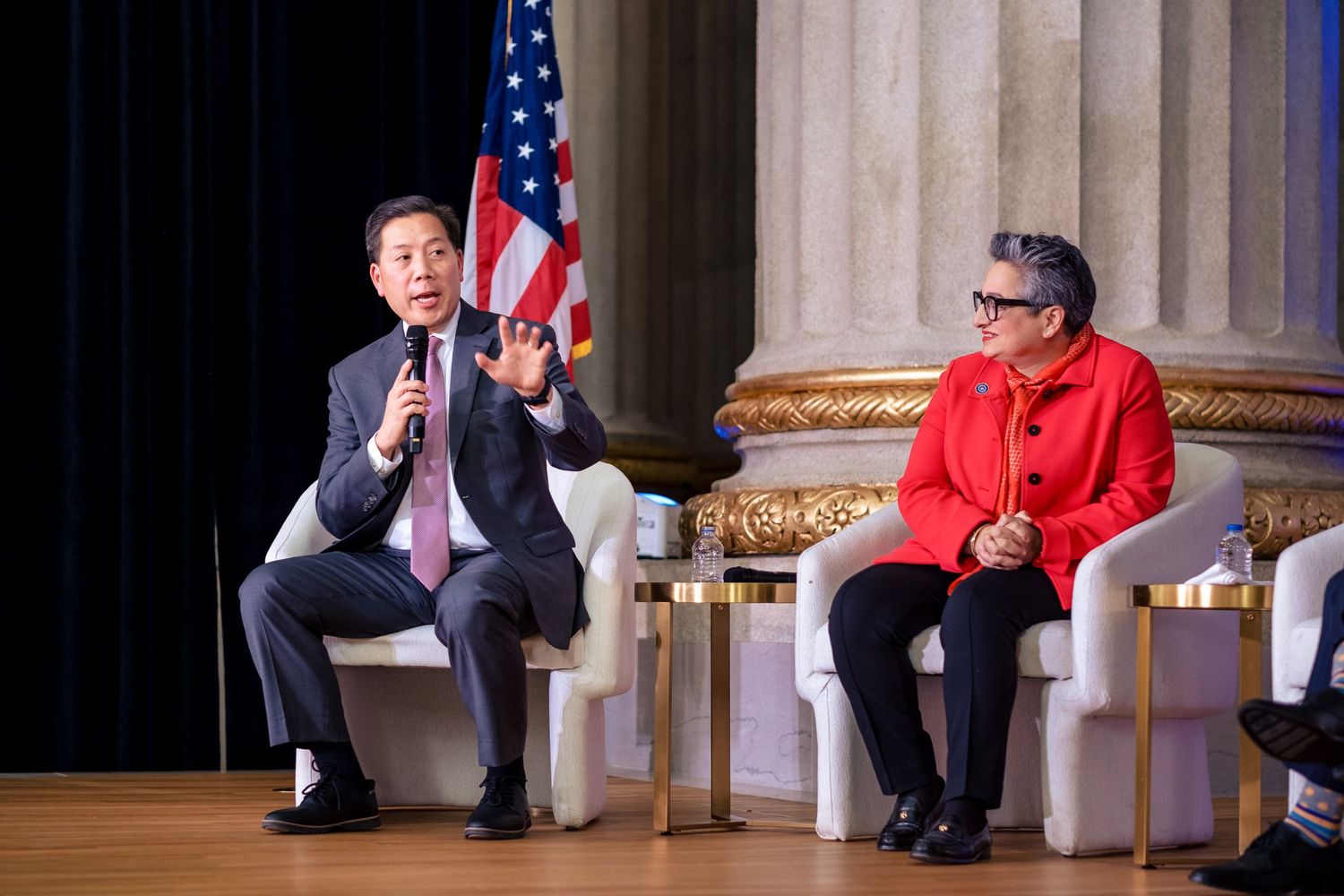 Two professionals speak on stage at a formal event with an American flag backdrop in an elegant gold venue.
