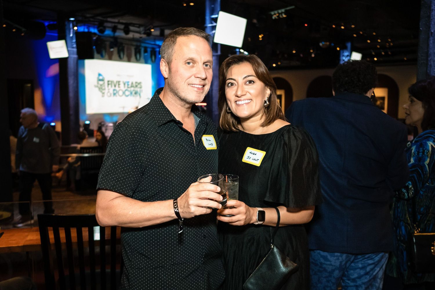 A man and woman smile together at a corporate anniversary party, holding drinks in a warmly lit event venue.