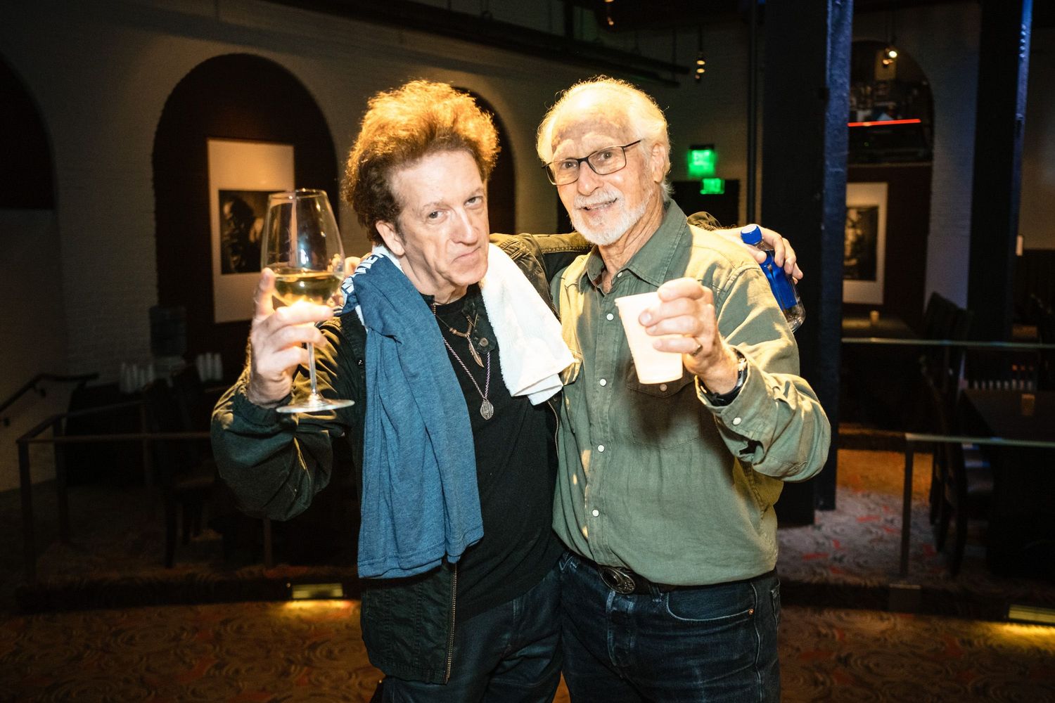 Two smiling men raise their drinks in a toast at an indoor venue, one holding wine and the other a cup.