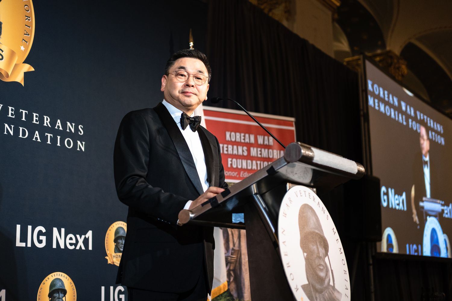 Man in tuxedo speaks at Korean War Veterans Memorial Foundation gala podium with LIG Nex1 sponsorship backdrop.