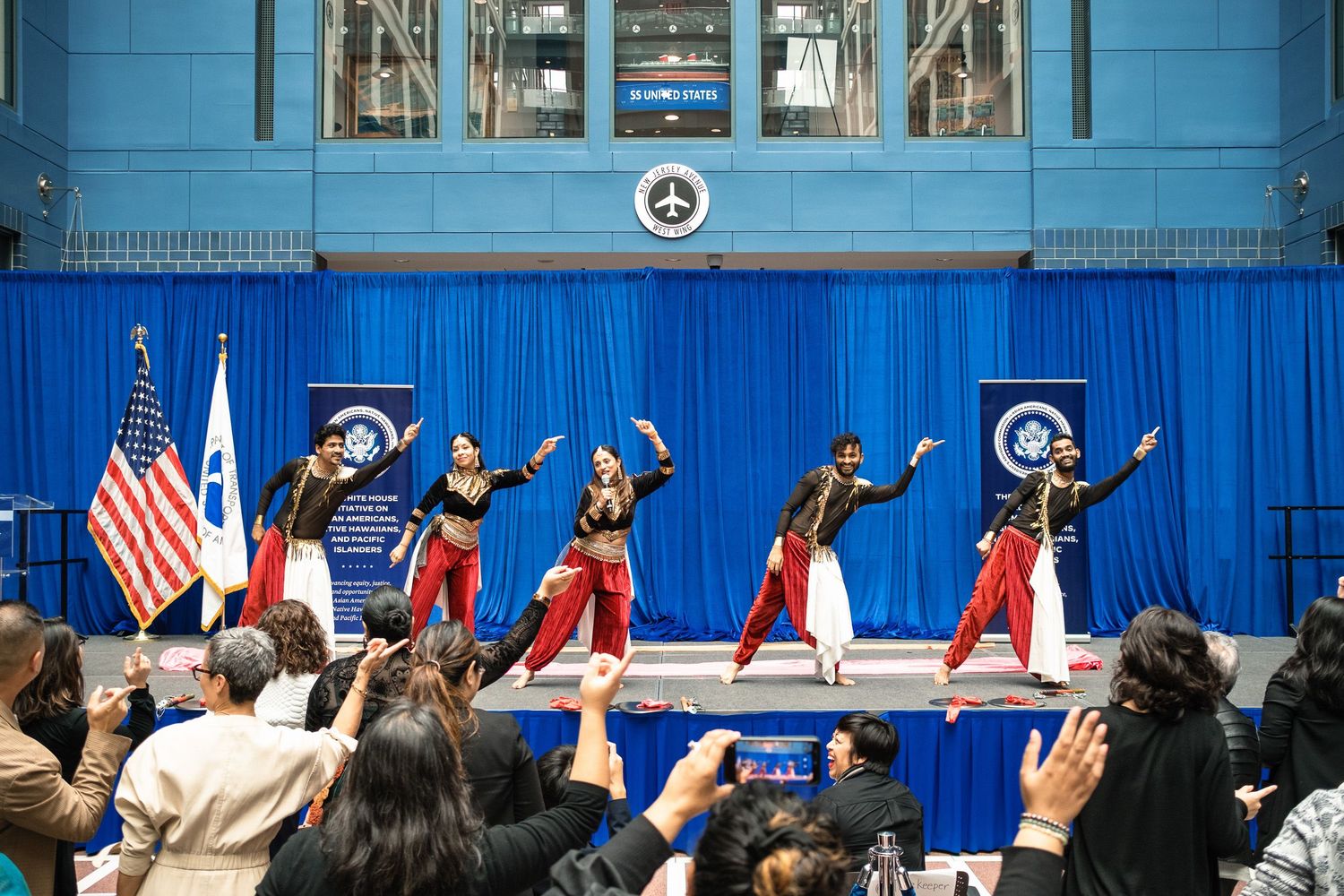 Colorful cultural dancers perform on stage before a crowd at a formal event with American flags and blue banners.