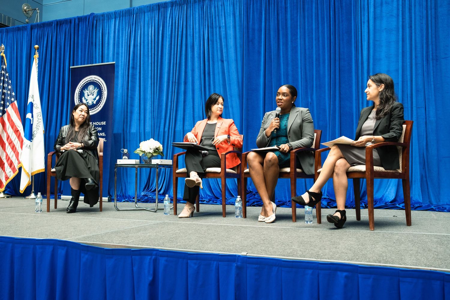 Four women seated on stage at a U.S. government panel discussion, with American and Israeli flags in the background.