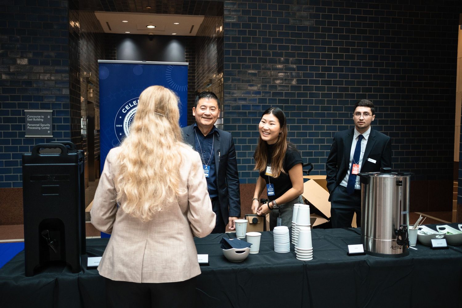 Professionals networking at a conference booth with coffee station, two hosts greeting a blonde attendee.
