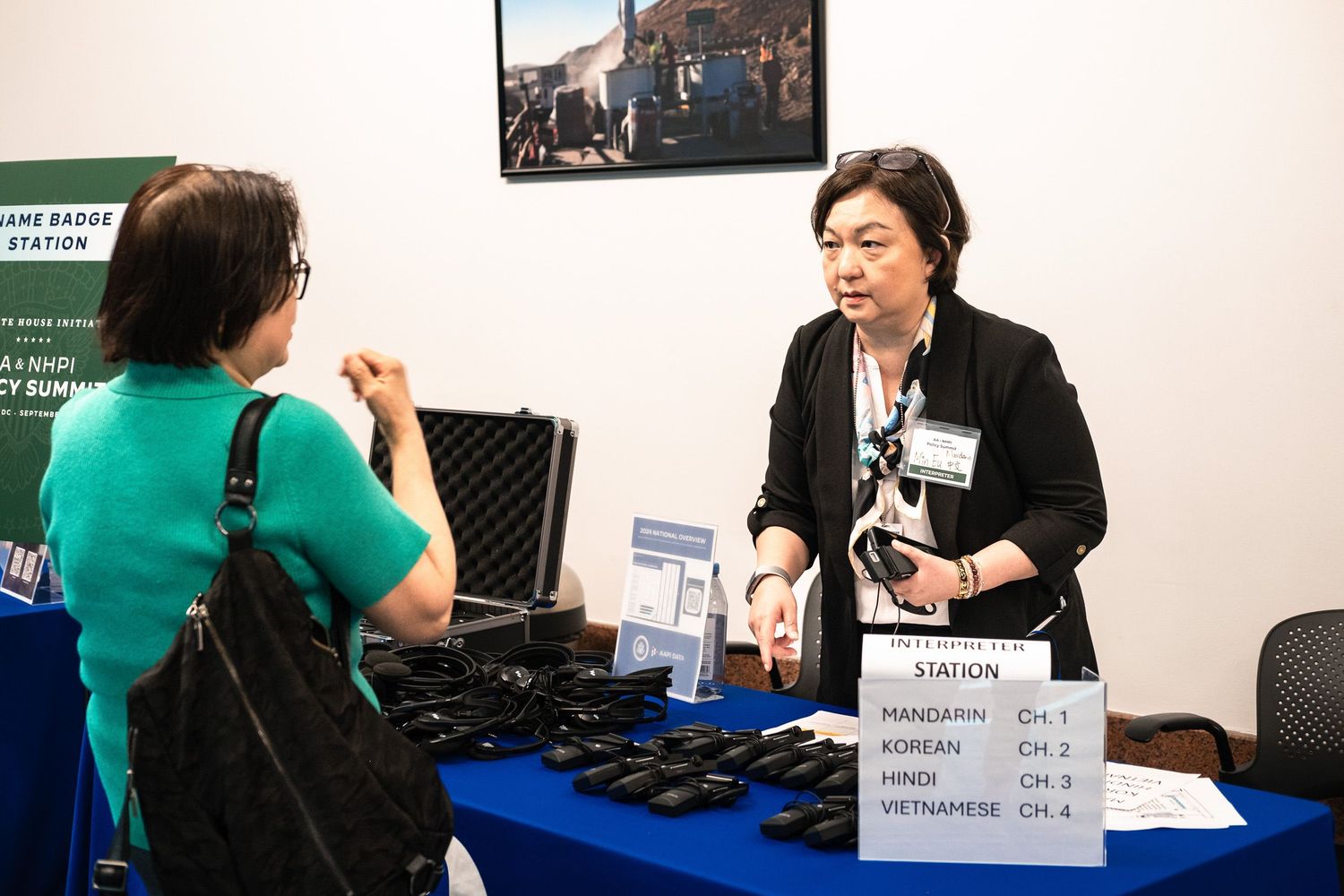 Two women at a multilingual translation station booth with radios for Mandarin, Korean, Hindi, and Vietnamese channels.