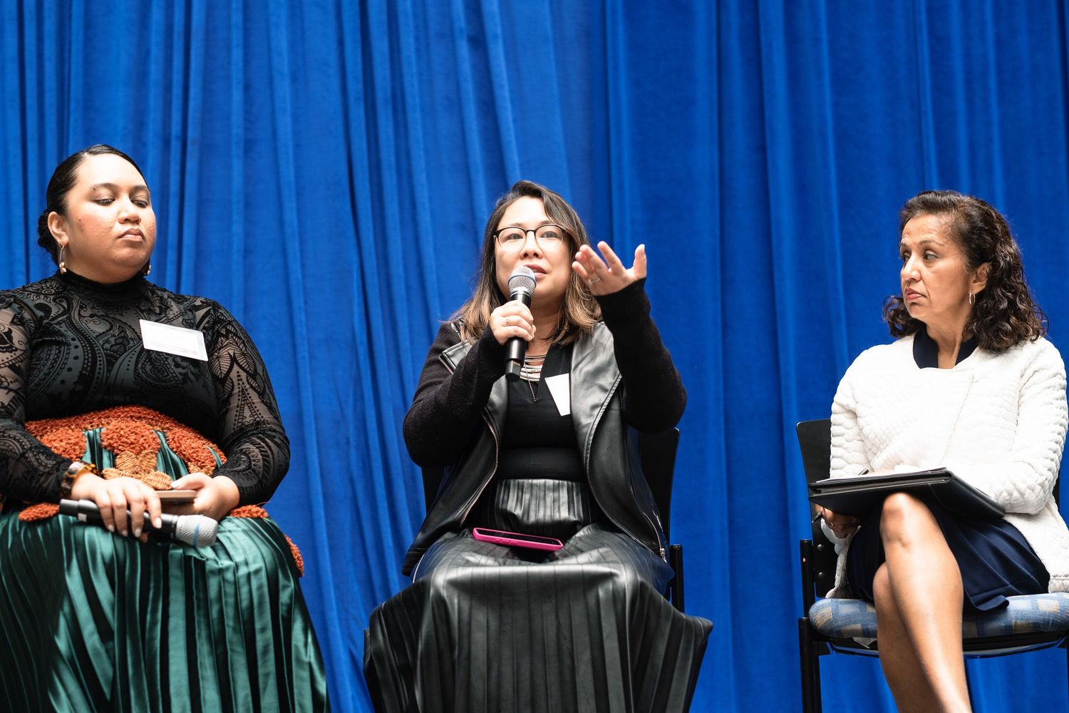 Three women sit on stage during a panel discussion, with one speaking into a microphone before a blue curtain backdrop.