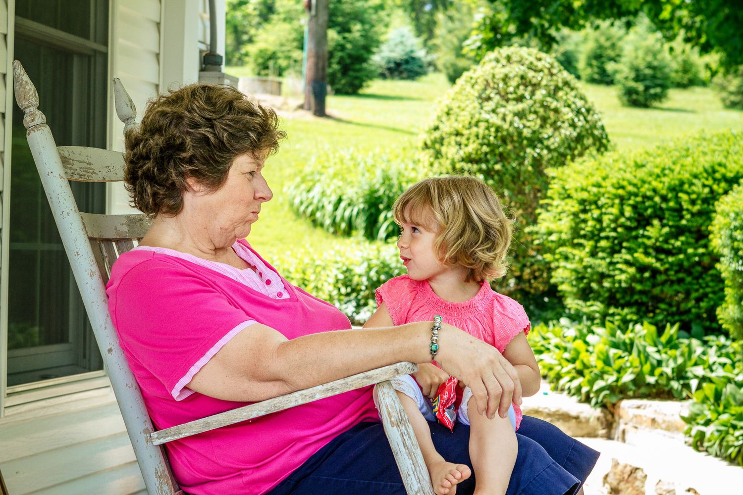 Grandmother and young granddaughter sharing a warm conversation while sitting on porch steps in matching pink shirts.