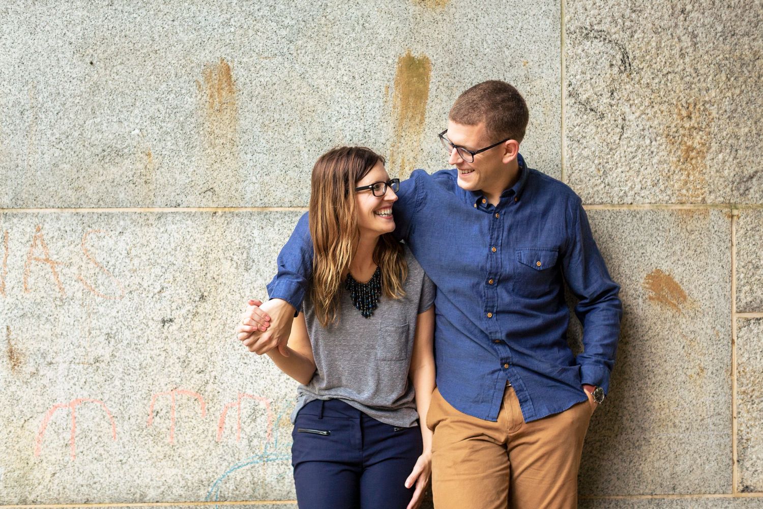 Smiling couple leaning against a stone wall, sharing a casual and affectionate moment together.