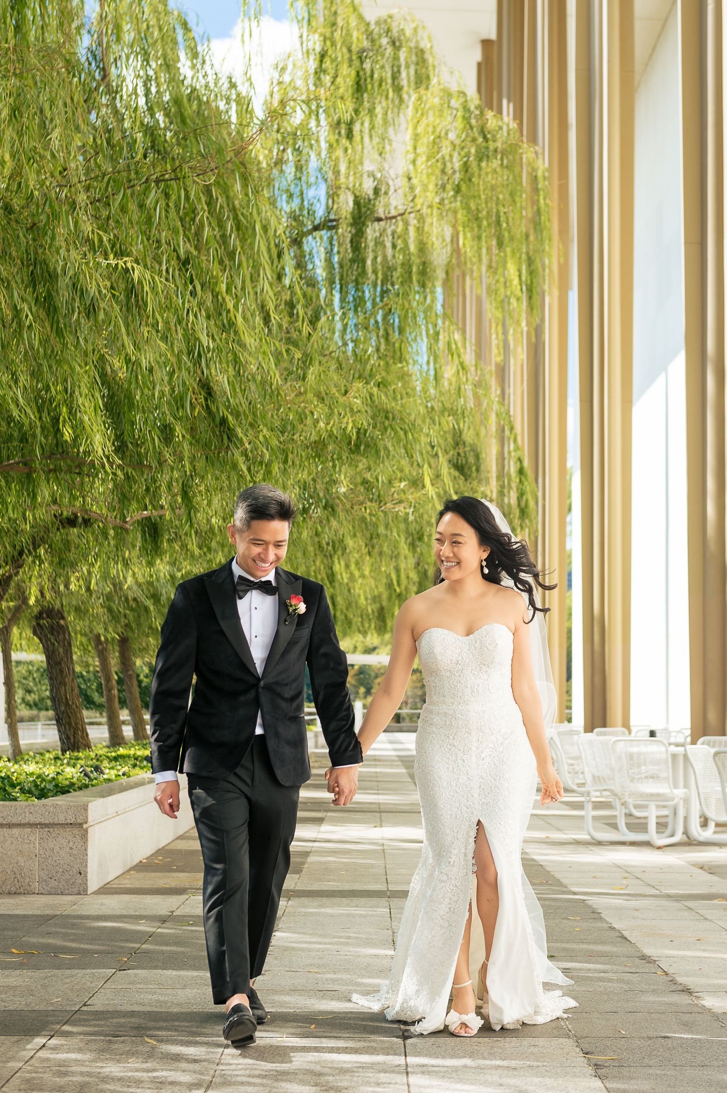 Elegant bride and groom walking hand in hand outdoors near weeping willow trees.