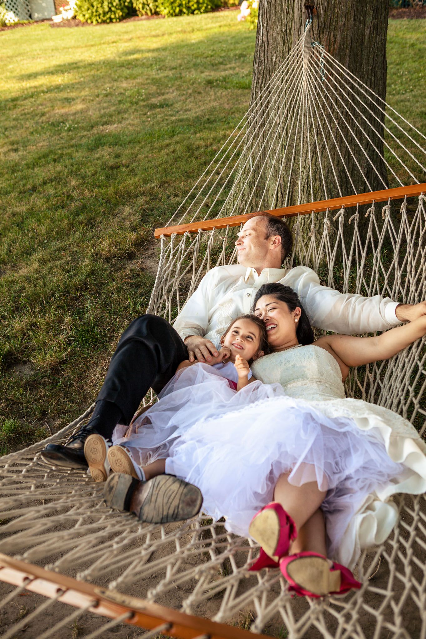Happy bride, groom, and child relax together in a hammock during outdoor wedding celebration.