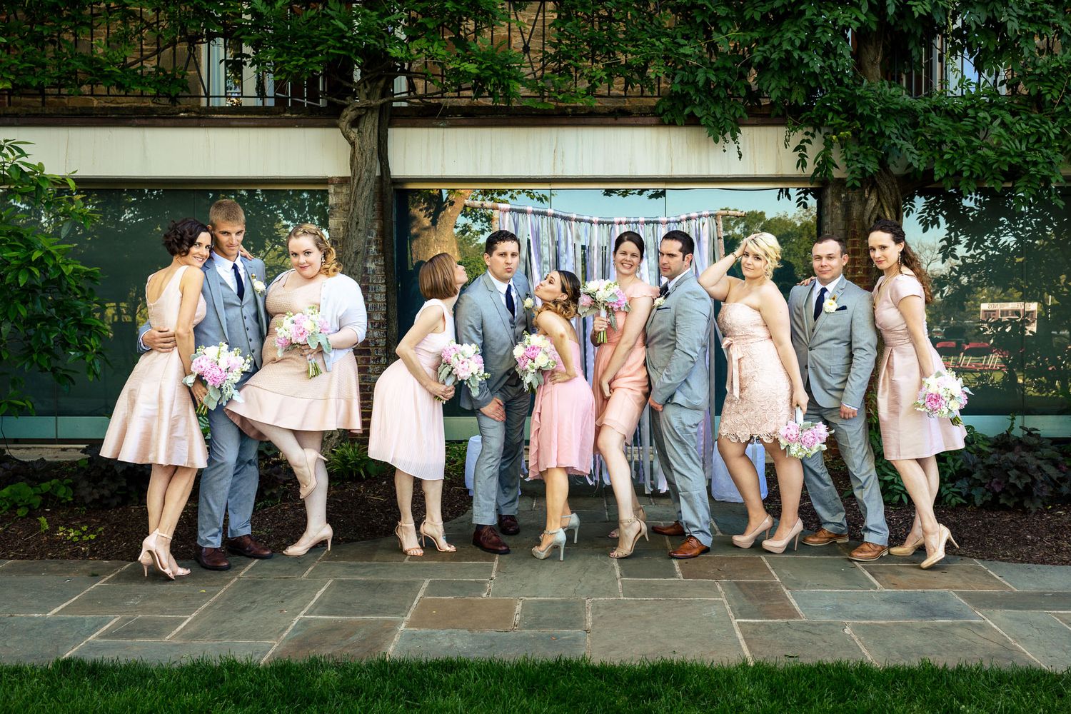 Wedding party in pink dresses and gray suits pose playfully on stone patio beneath leafy vines.