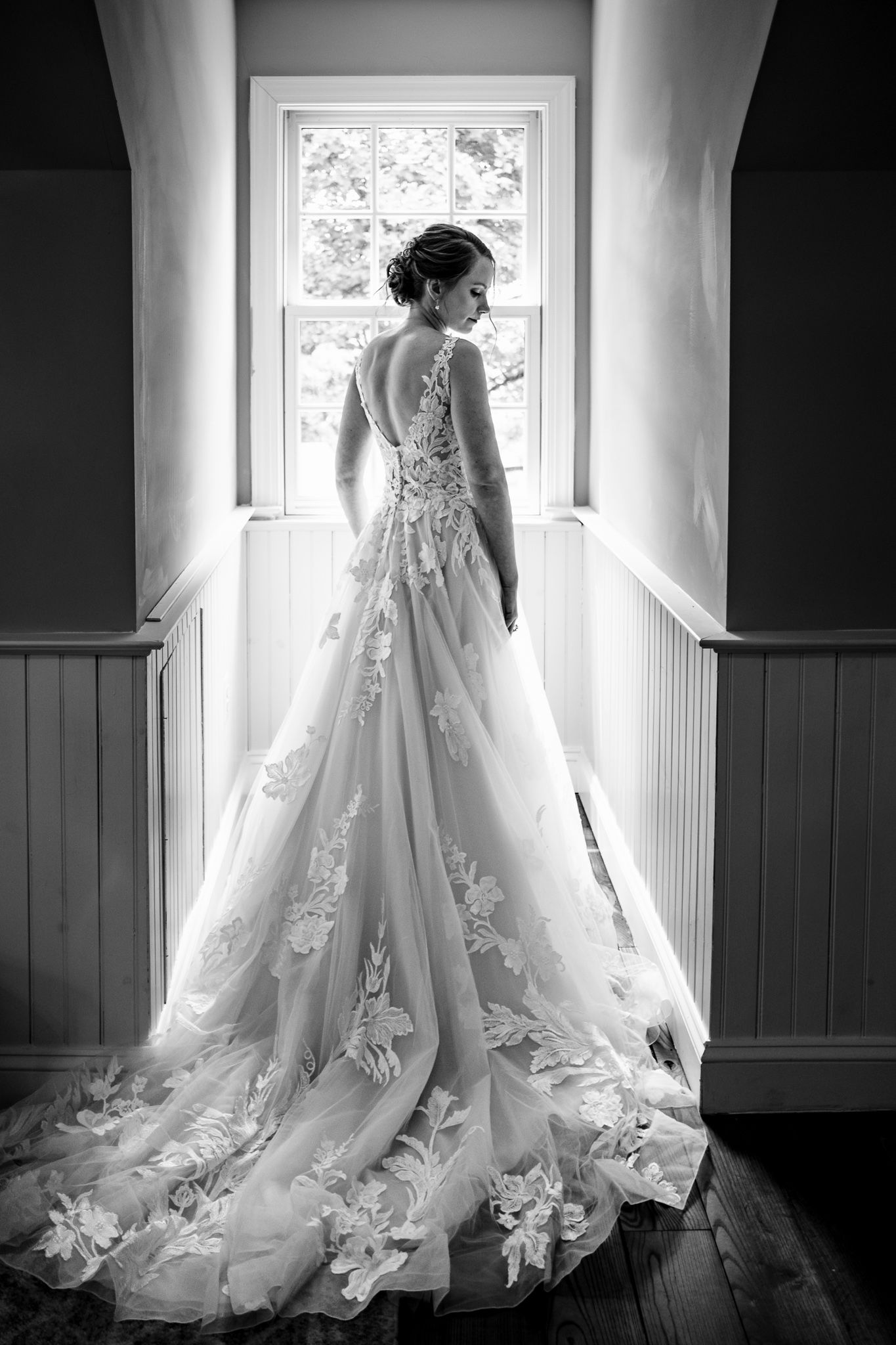 Black and white photo of bride in floral lace ballgown standing before window.
