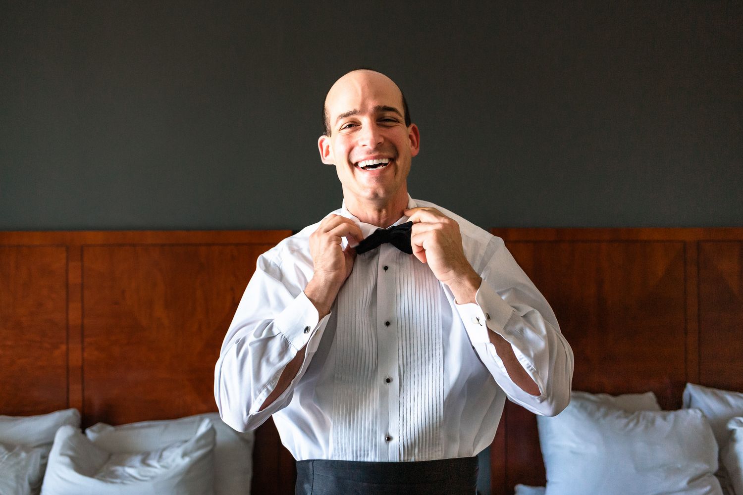 Smiling bald groom adjusts his black bow tie while getting ready for his wedding day.