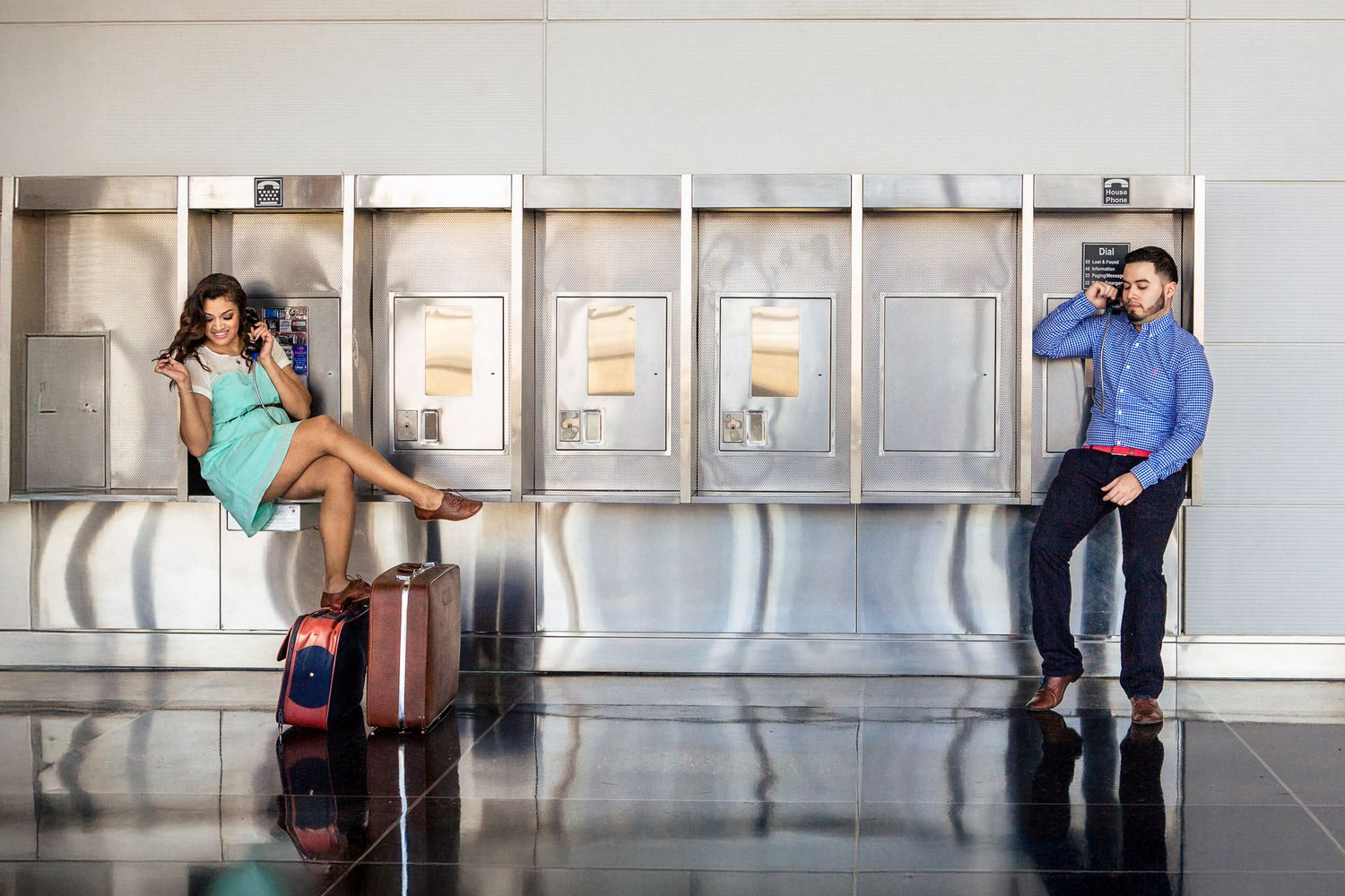 Two travelers use payphones at an airport terminal, sitting on luggage while waiting for their flight.