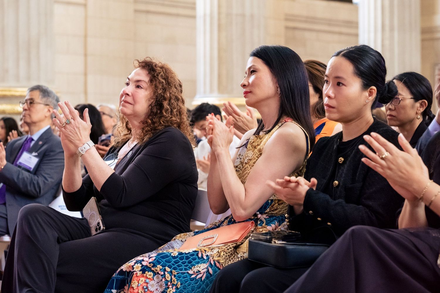 Three women applauding at a formal multicultural event in a grand venue.