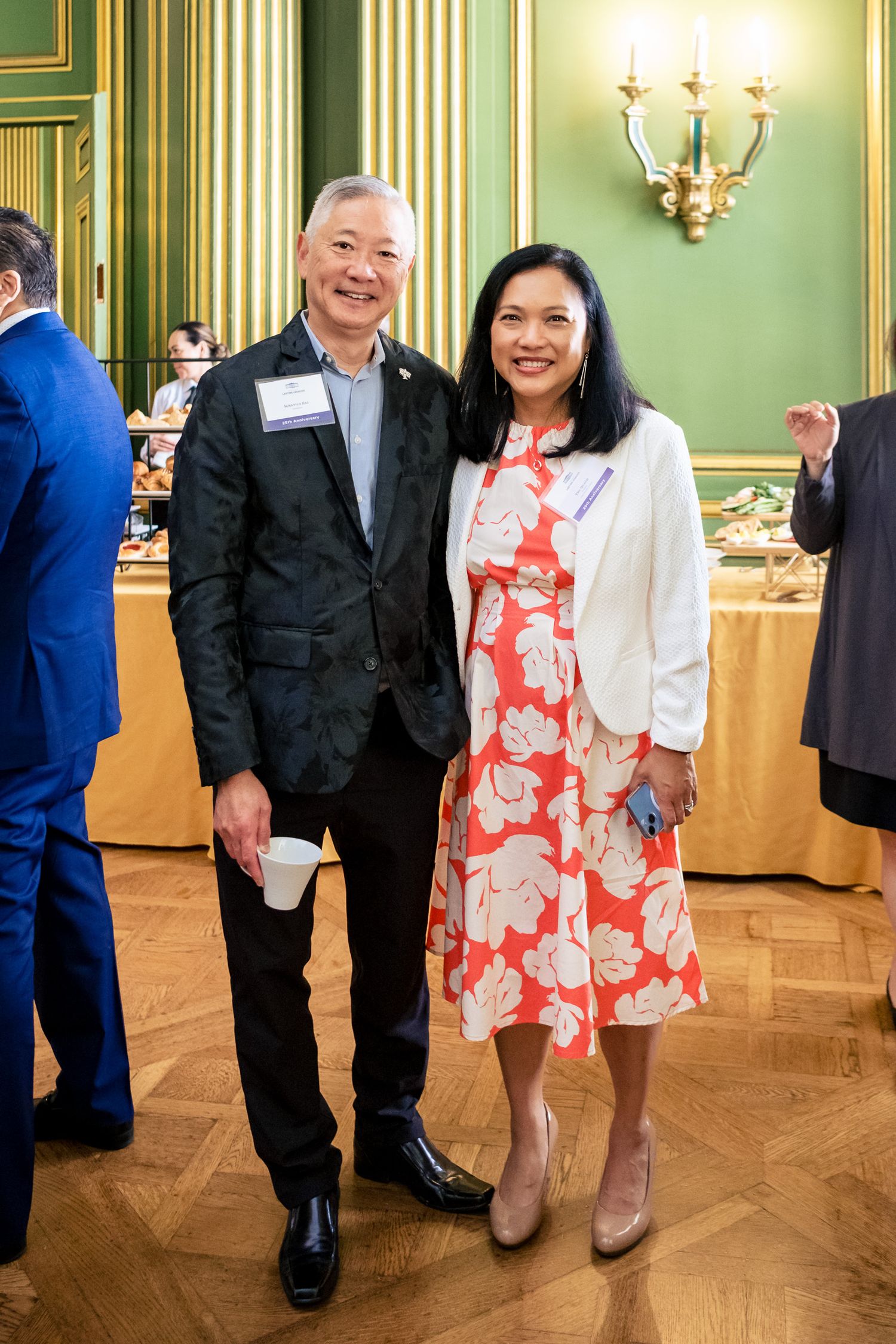 Two smiling attendees pose together at an elegant formal event in a grand ballroom with green walls.