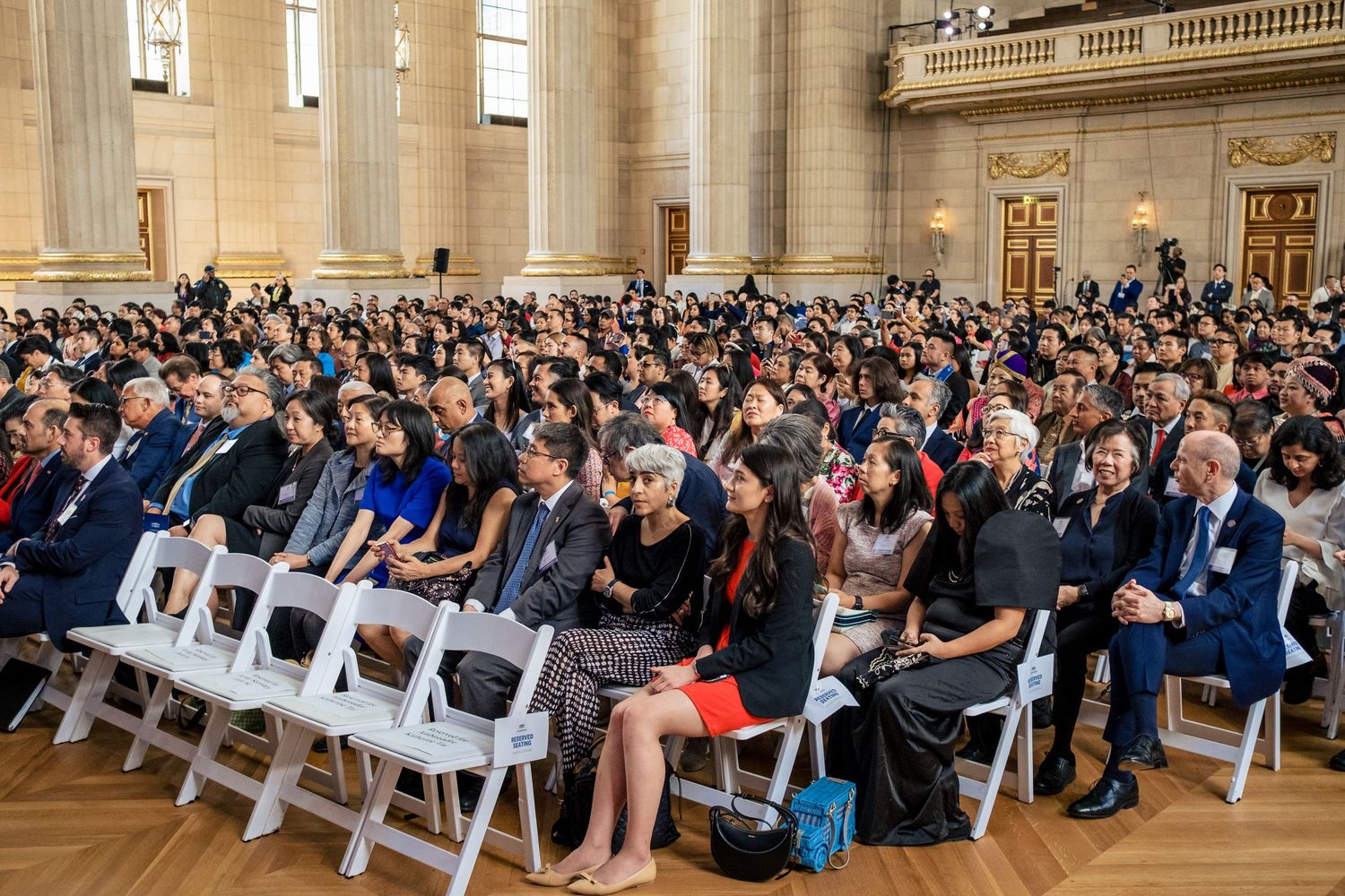 Large diverse audience seated in rows of white chairs at a formal indoor ceremony in a grand hall.