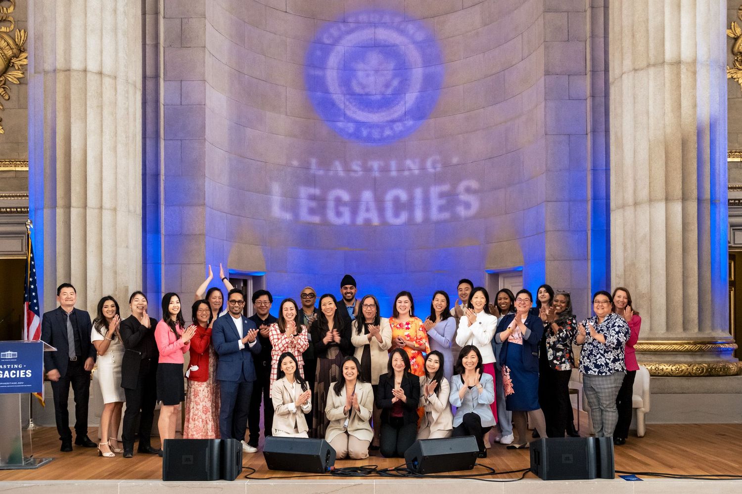 Group photo at Lasting Legacies event inside ornate hall with blue lighting, diverse attendees smiling and applauding together.