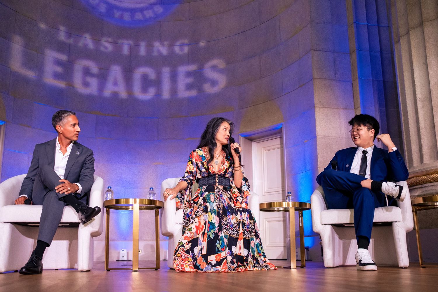 Three panelists seated on stage at a Lasting Legacies event, with a woman in floral dress as moderator.