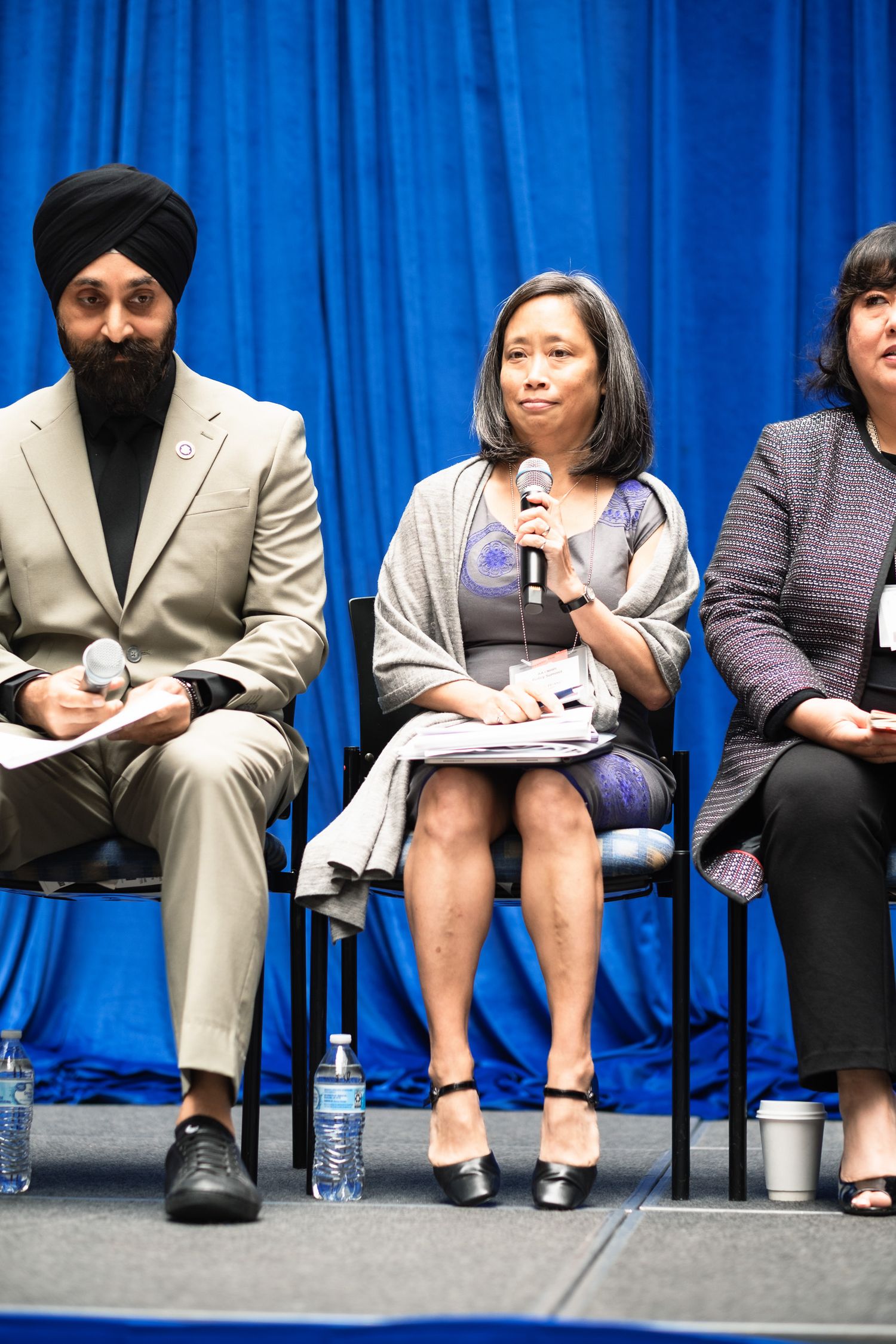 Three panelists seated on stage at a formal event, with a blue curtain backdrop, one speaking into a microphone.