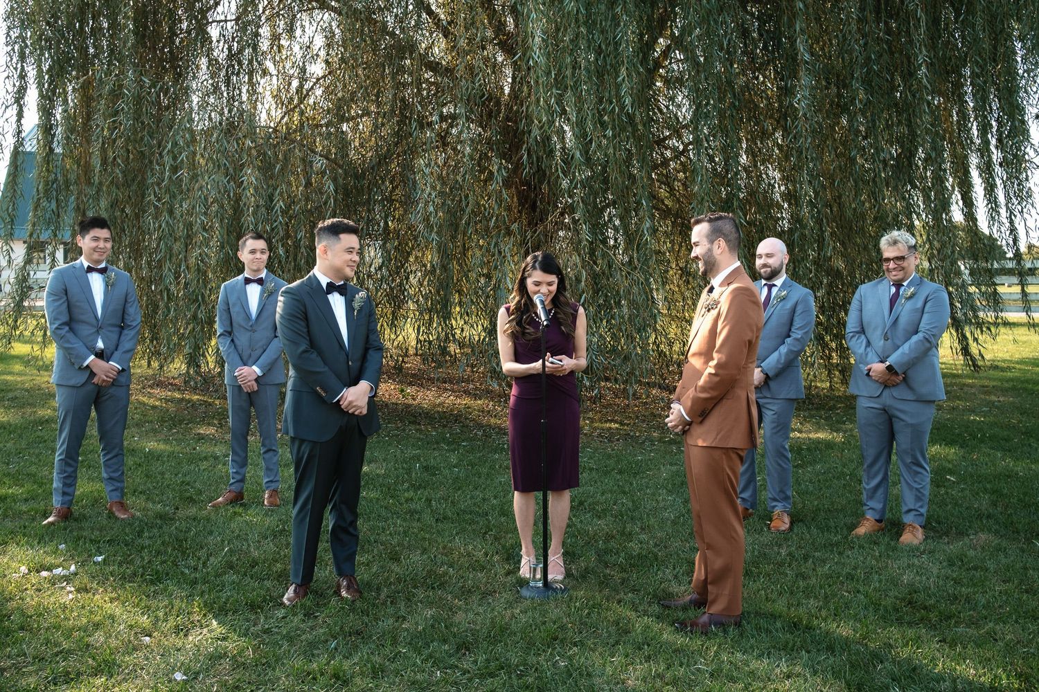 Two grooms stand before their officiant with groomsmen during an outdoor wedding ceremony by a willow tree.