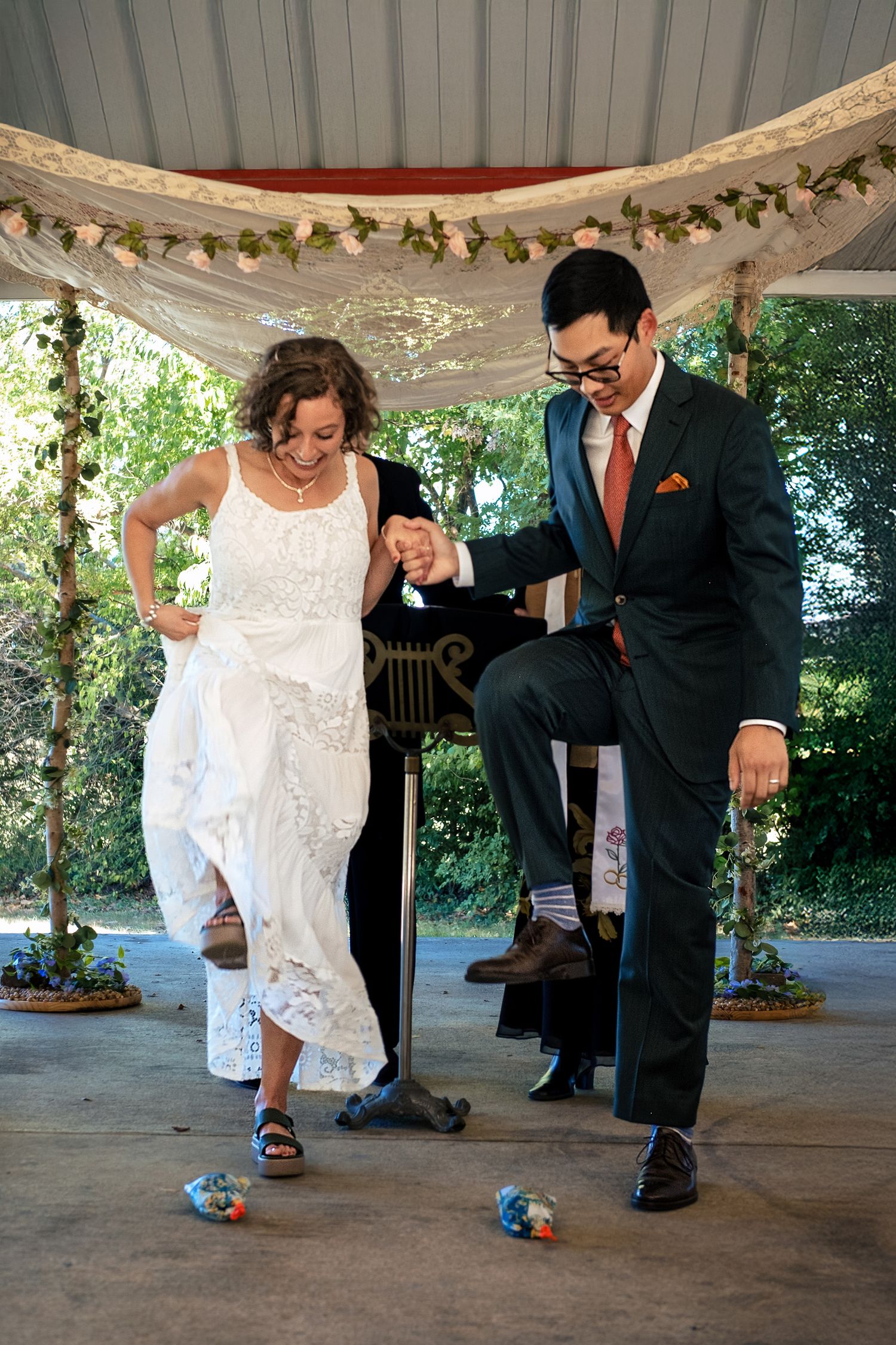 Bride and groom stomping on glass during outdoor Jewish wedding ceremony under floral chuppah.
