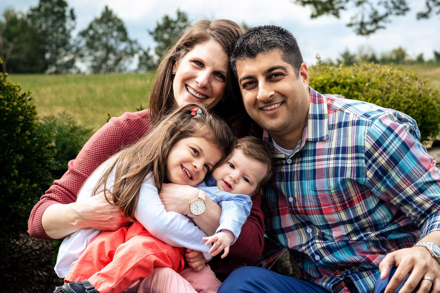 Happy family of four parents and two children embracing outdoors in green park setting.