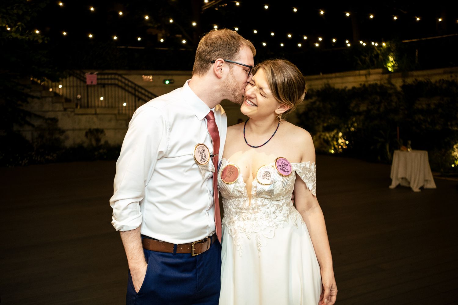 Groom kissing bride on the cheek at their wedding reception under warm evening lights.