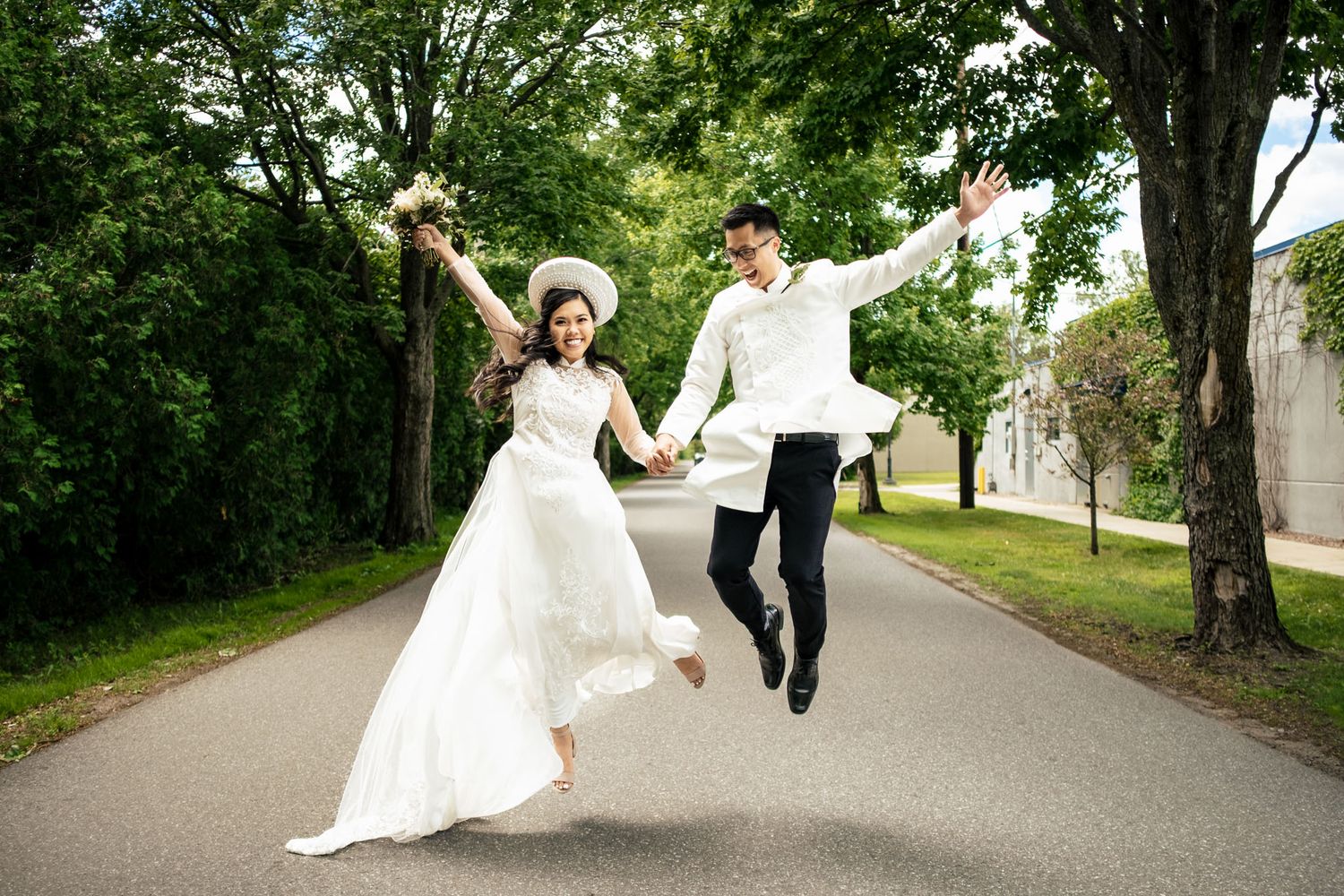 Joyful bride and groom jump together hand-in-hand on a tree-lined path in white attire.