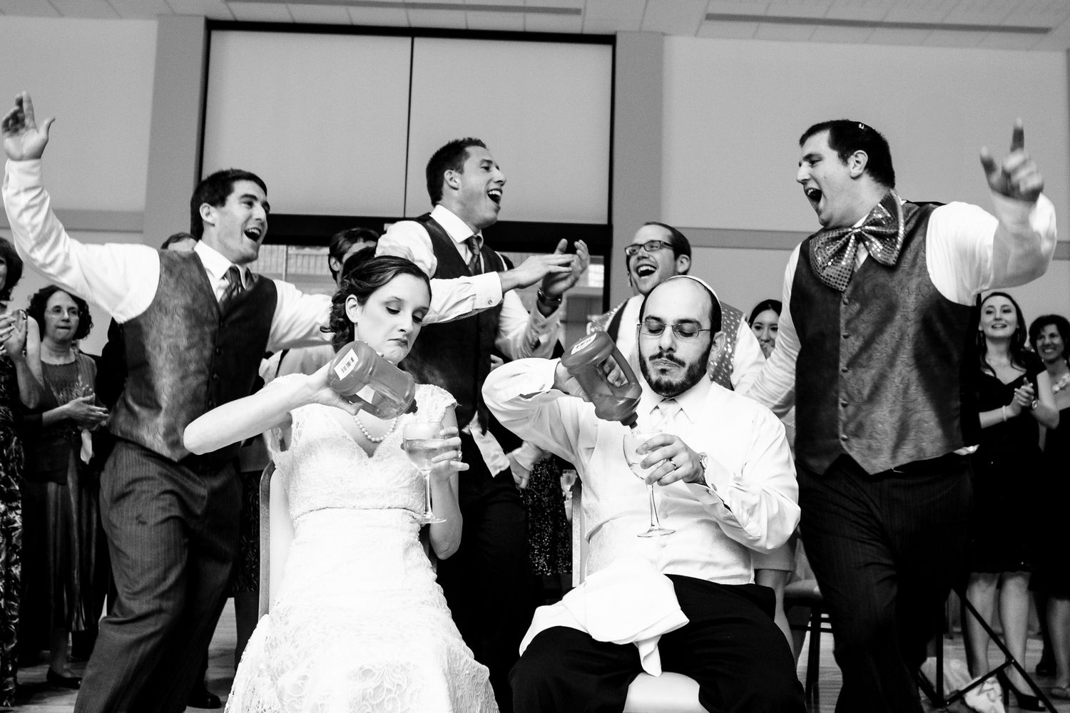 Wedding party celebrates joyfully on dance floor with bride and groom in black and white photo.