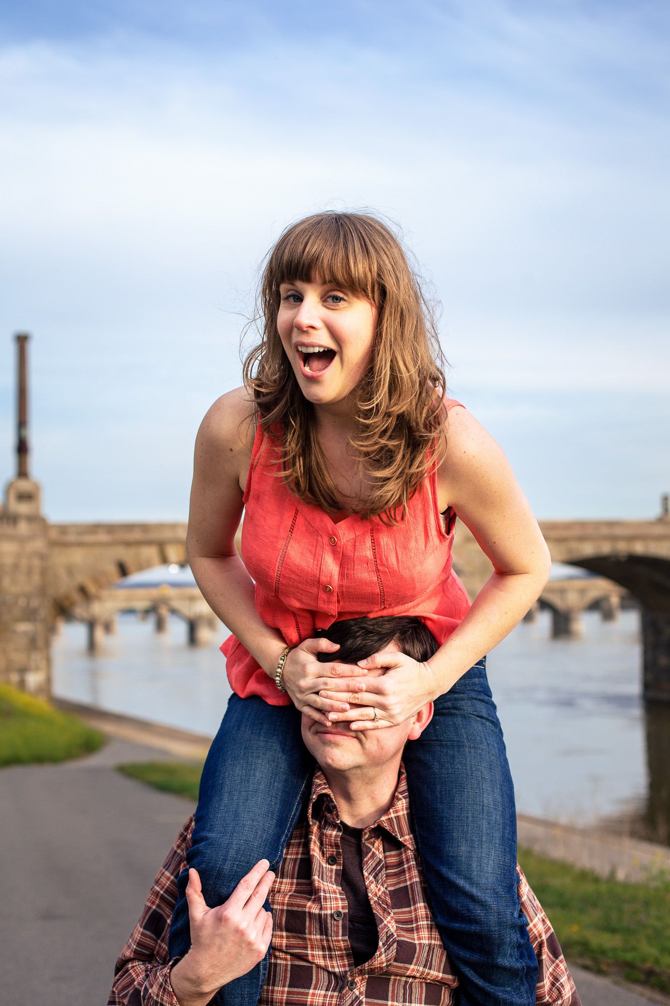 Excited woman sitting on mans shoulders covering his eyes near a river bridge outdoors.