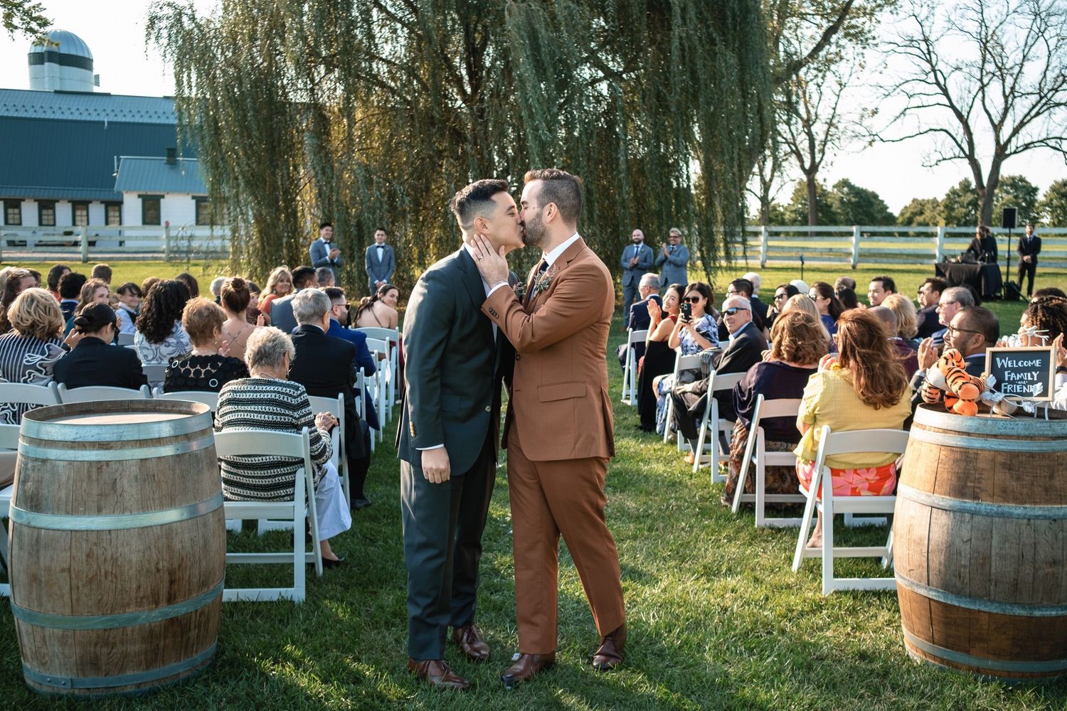 Newlywed same-sex couple shares a kiss at the altar with guests seated along the aisle at outdoor wedding.