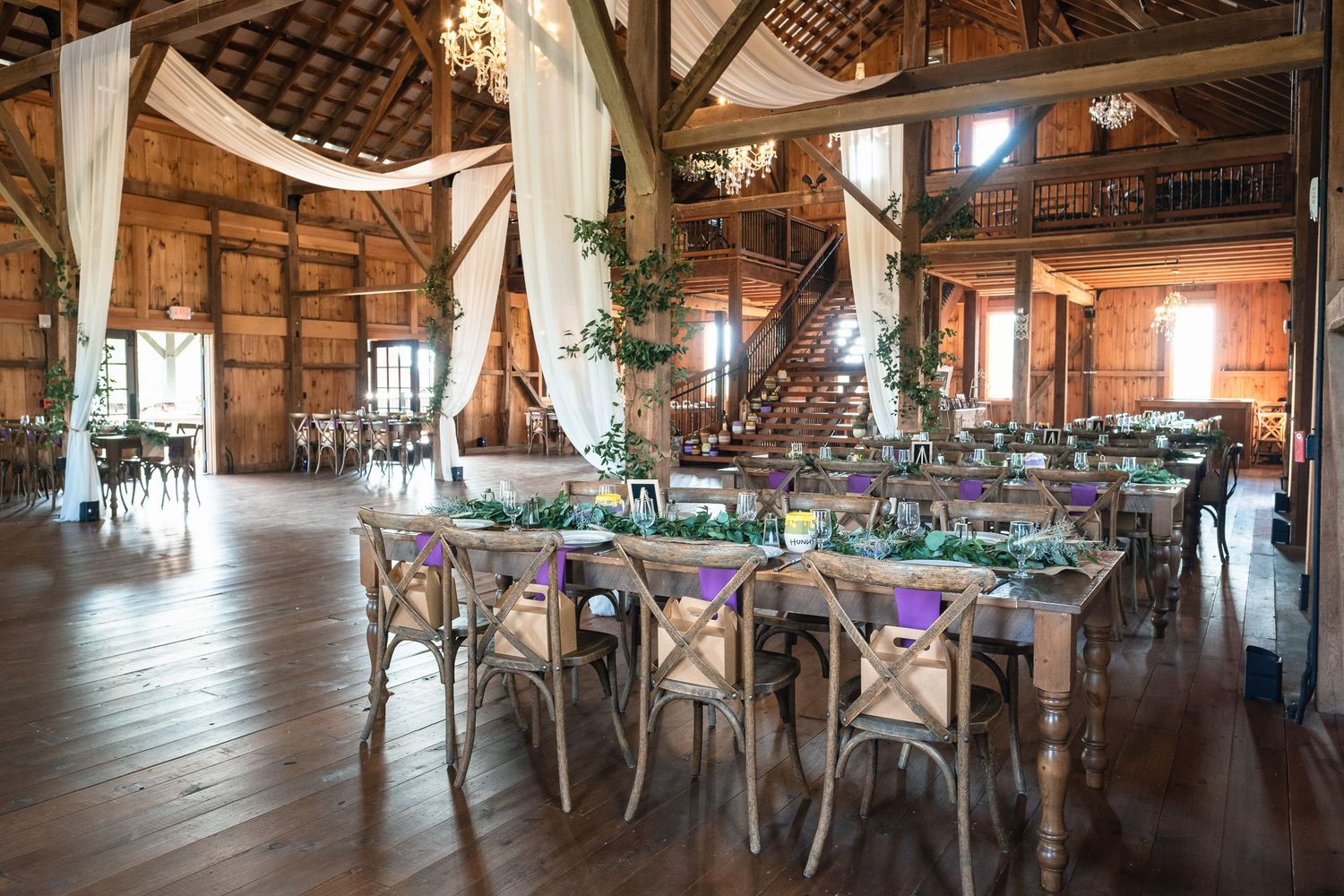 Elegant rustic barn wedding reception with long tables, purple accents, and draped white fabric decor.