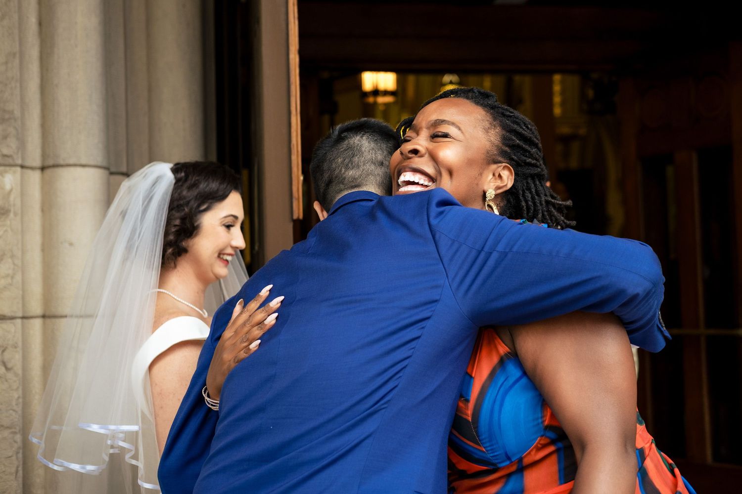 Joyful wedding guest hugging groom in blue suit while bride in white gown and veil smiles nearby at church entrance.