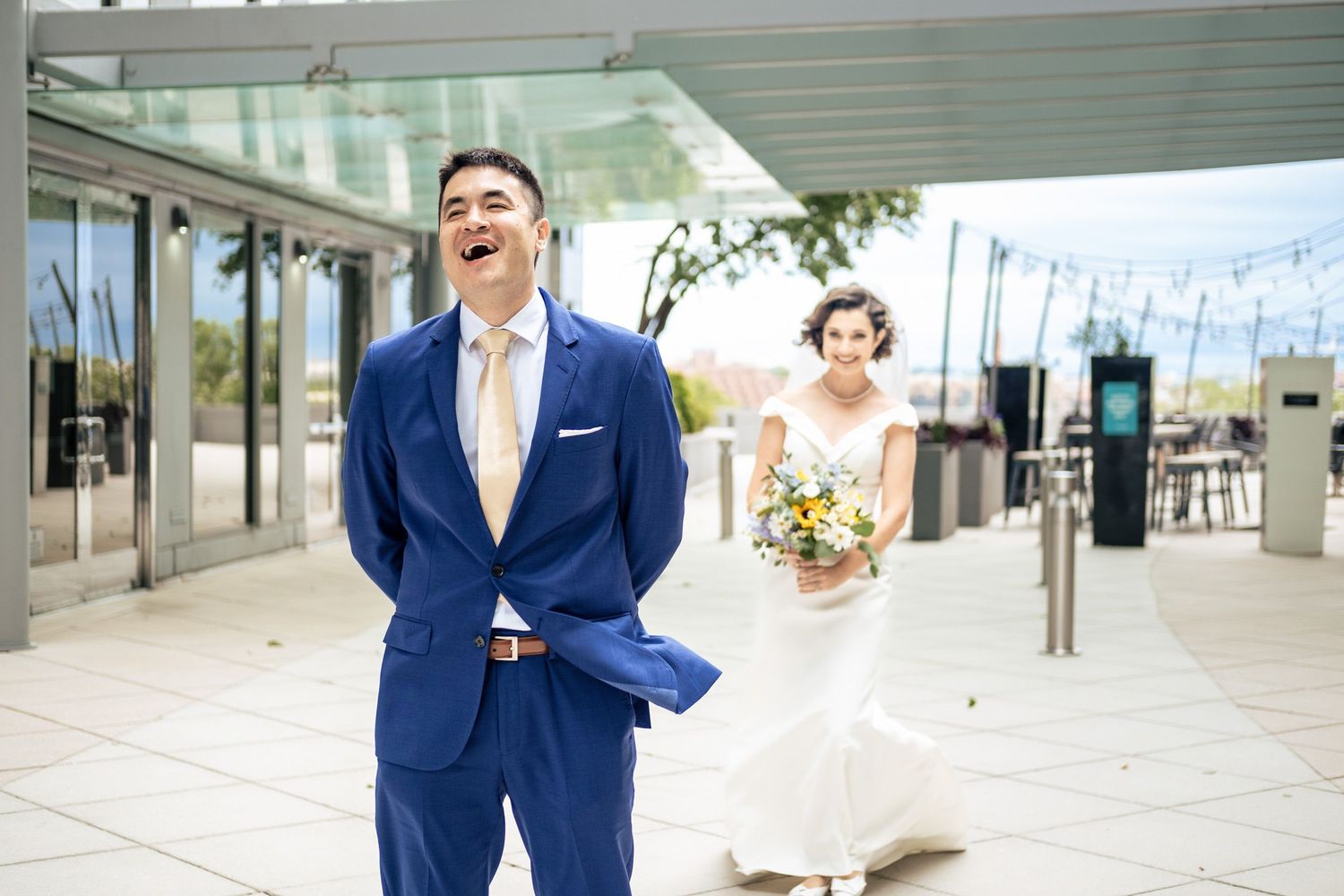 Groom in blue suit laughs joyfully during first look as bride in white gown approaches with yellow bouquet.