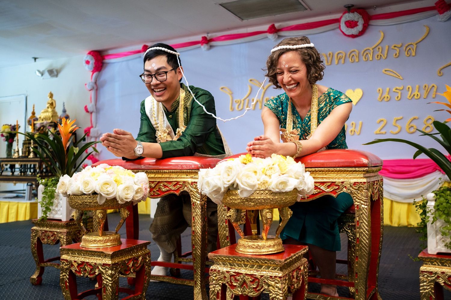 Couple in traditional Thai wedding attire laughing joyfully during a Buddhist blessing ceremony.