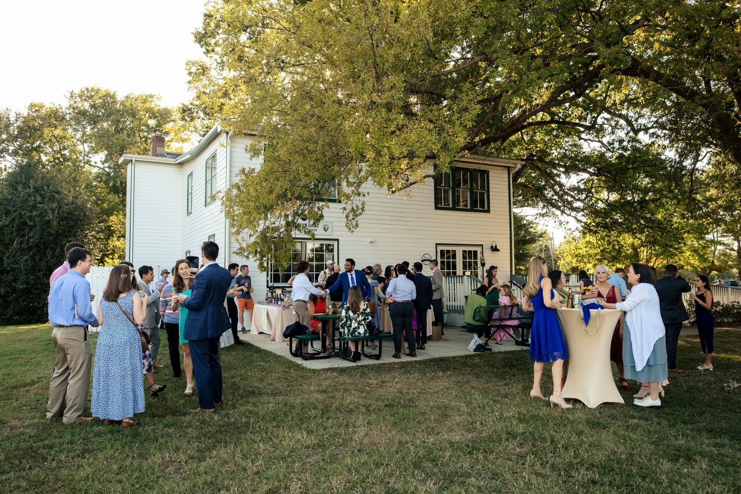 Wedding guests mingling on a sunny lawn outside a white two-story house beneath a large shade tree.