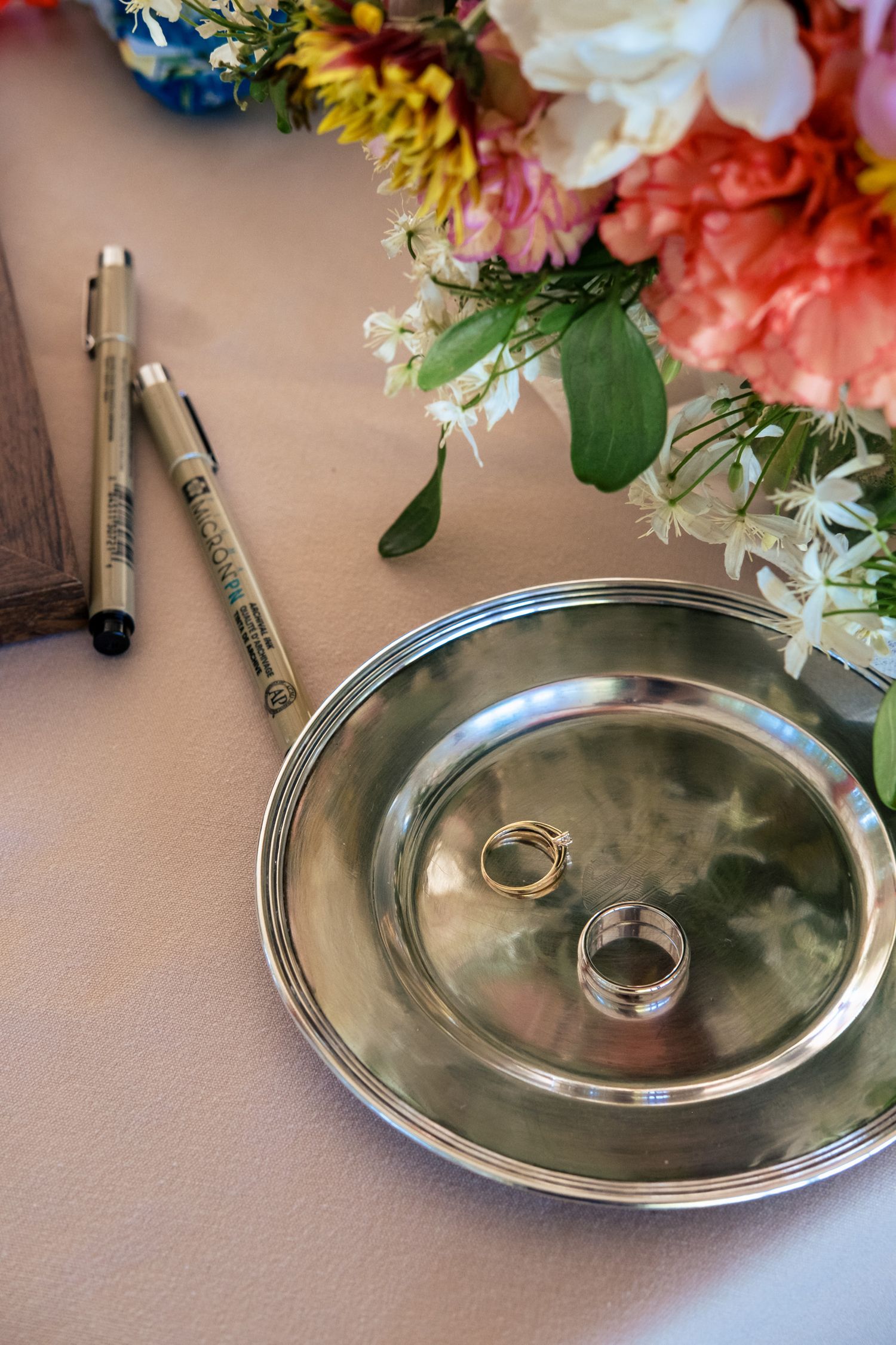 Two gold wedding rings rest on a silver dish beside pens and a colorful floral bouquet on a table.