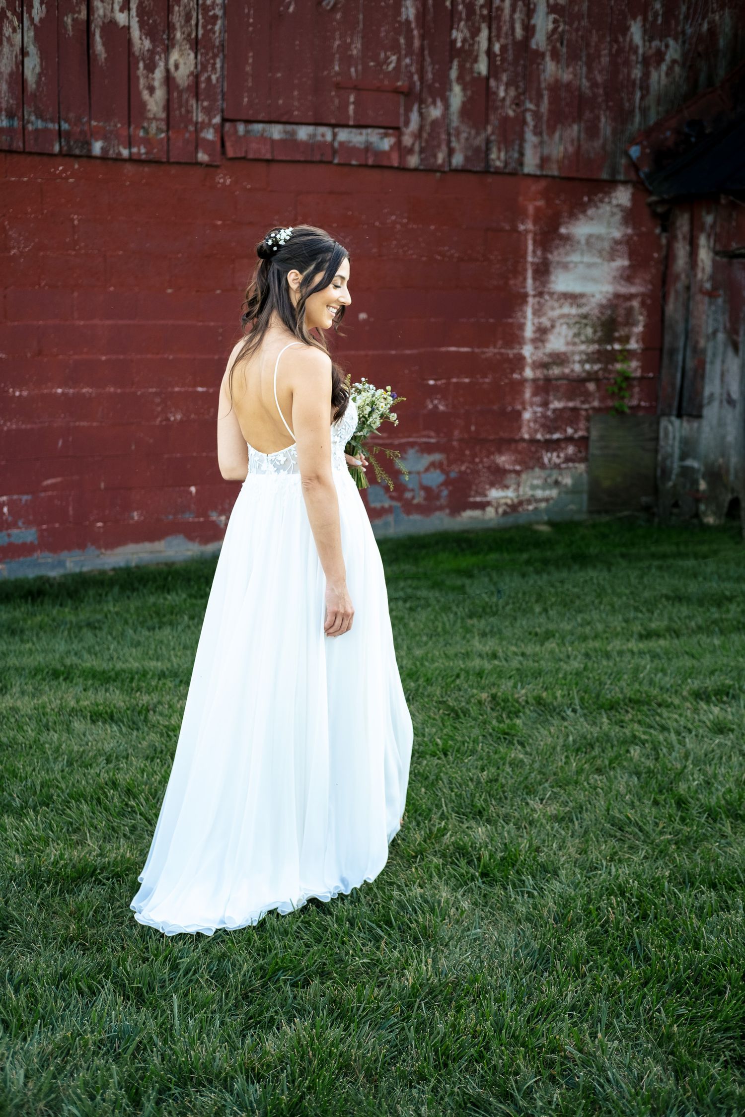 Bride in a backless white gown holds a bouquet while standing on grass near a red barn.