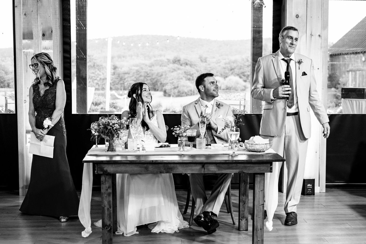 Black and white photo of groom giving wedding speech while bride sits at sweetheart table.