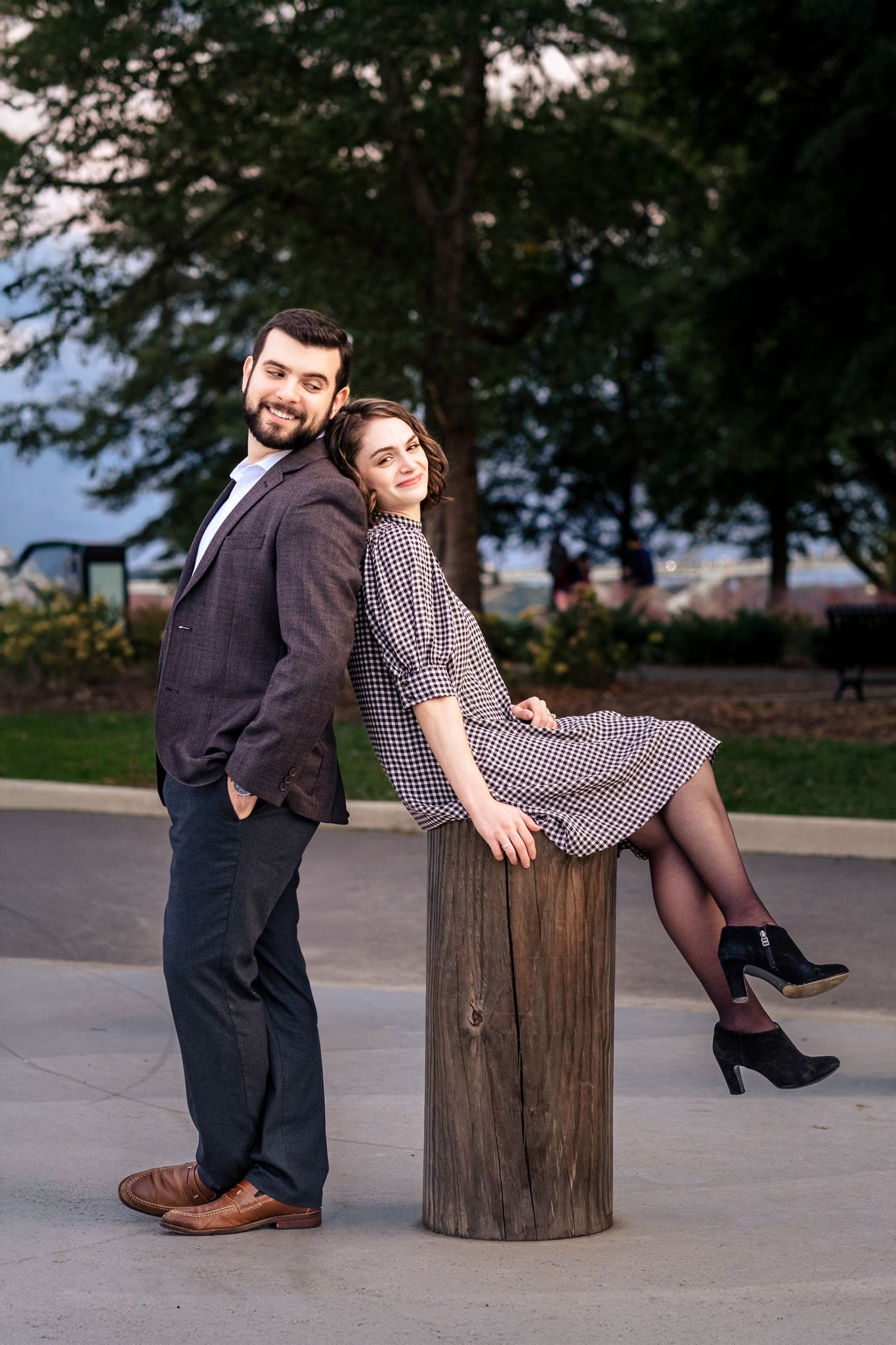 Smiling couple posing back-to-back near a wooden post in a park at sunset.