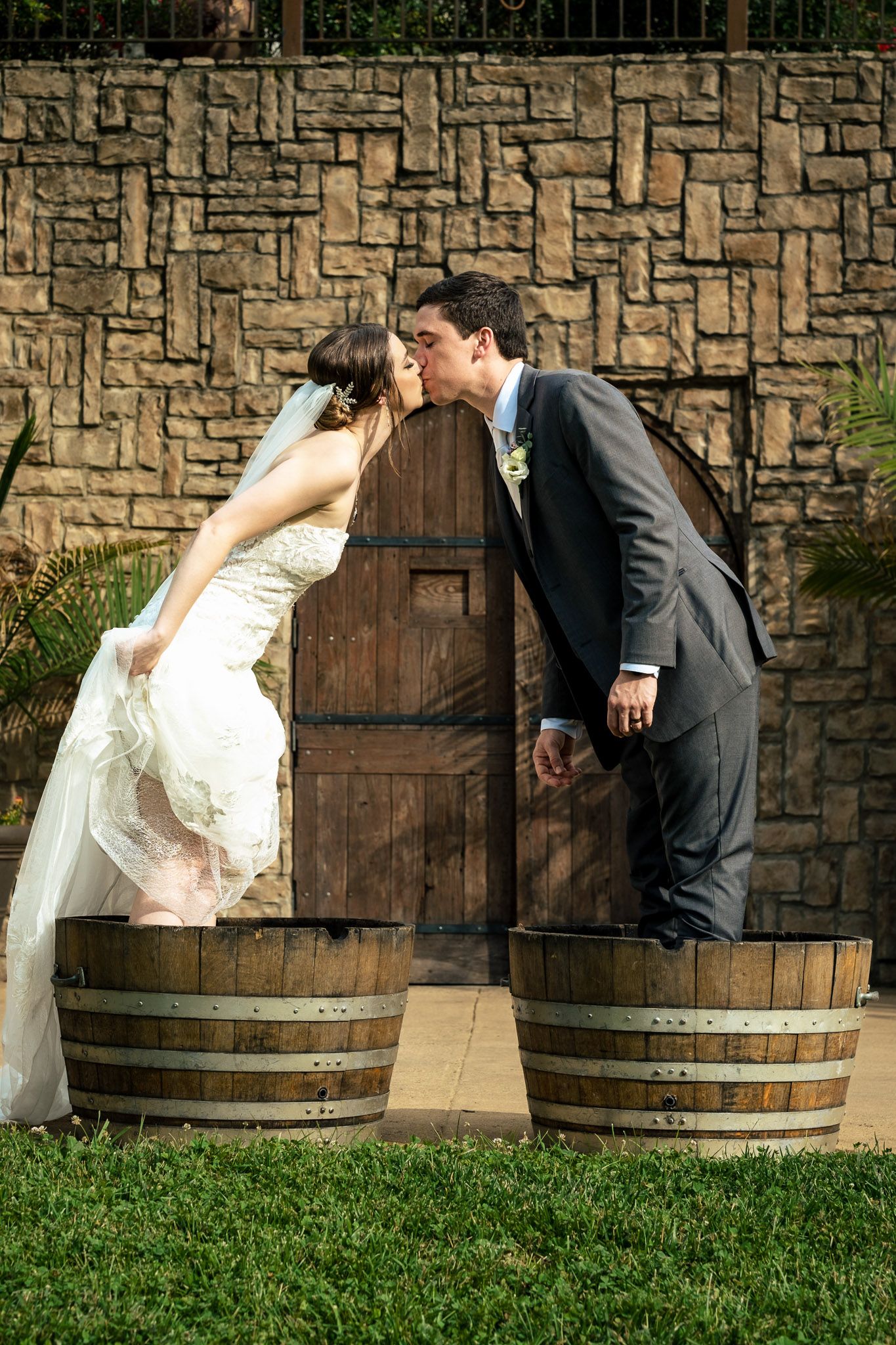 Bride and groom share a romantic kiss while standing in wooden wine barrels before a rustic stone wall.