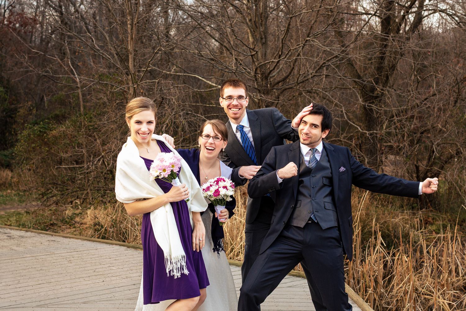 Wedding party of four poses playfully on a wooden bridge in an outdoor setting.