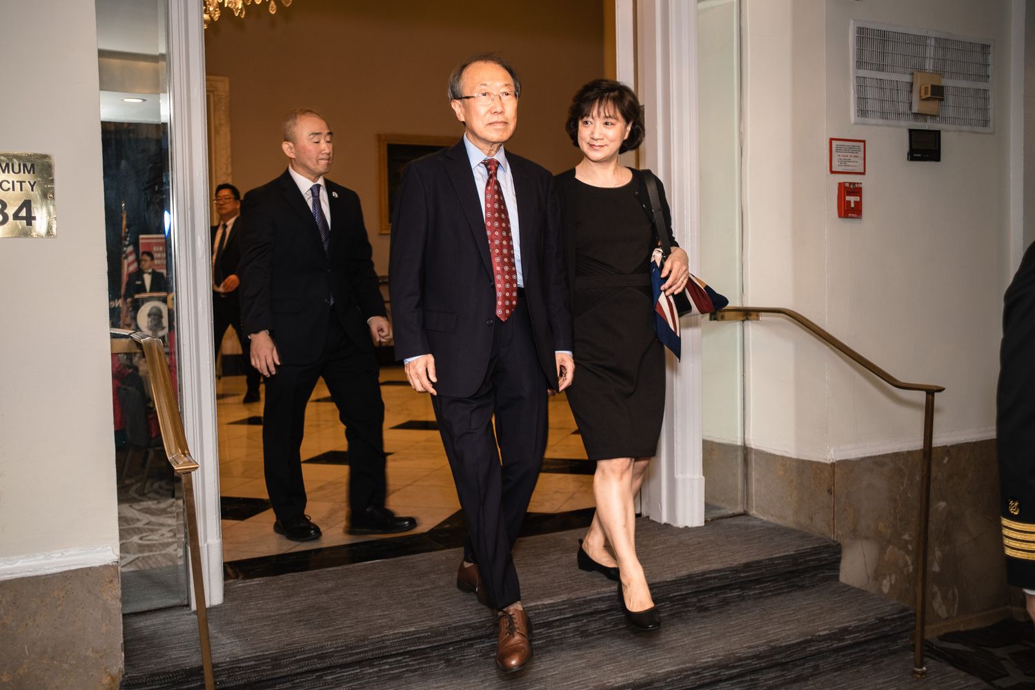 Two professionals in formal attire descend steps in an elegant building, with a suited man visible in the background.
