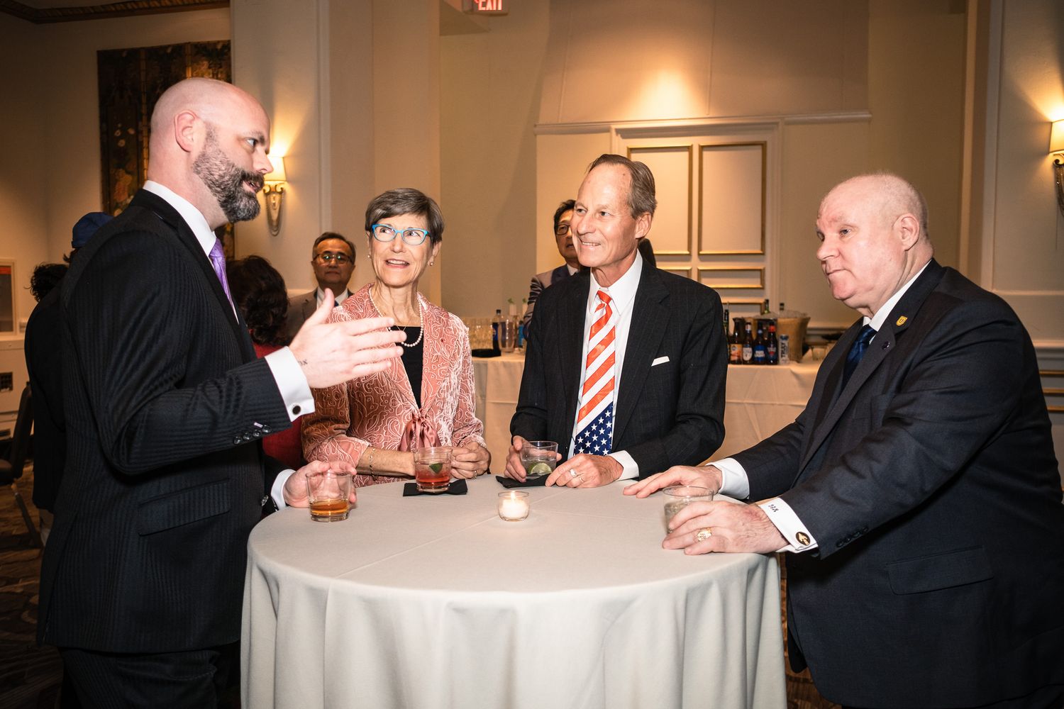 Five formally dressed professionals socializing around a round table at an upscale networking event.