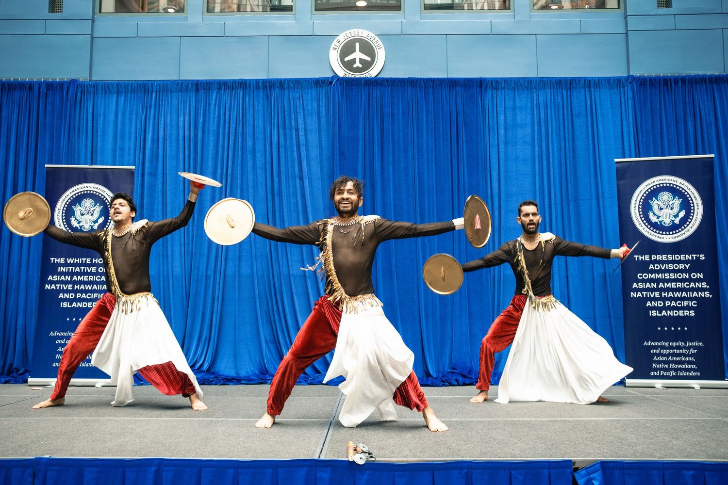 Three performers in traditional attire dance with cymbals on a blue-draped stage at a White House Asian American event.